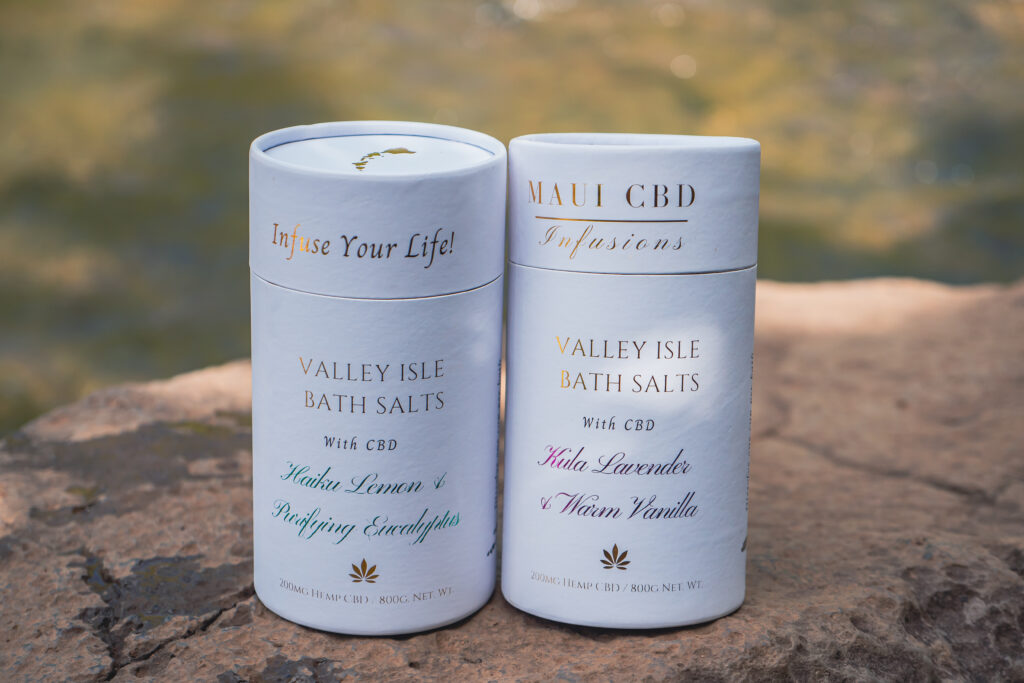 Two containers of Valley Isle Bath Salts with CBD: Haiku Lemon & Purifying Eucalyptus and Kula Lavender & Warm Vanilla, displayed outdoors on a natural stone surface