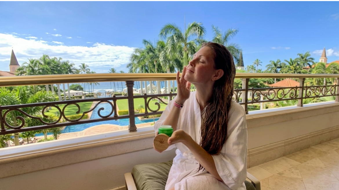 A woman in a white bathrobe sits on a tropical resort balcony overlooking a pool and the ocean, applying Maui CBD Infusions face cream from a green jar with a bamboo lid.
