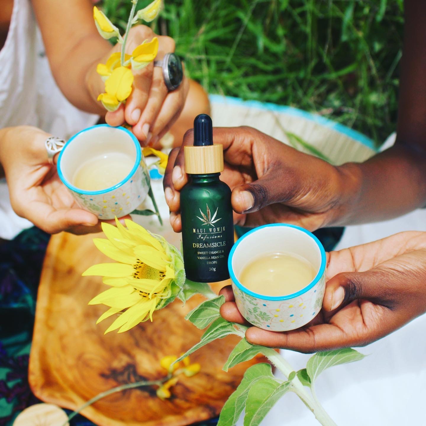 Two people holding small tea cups next to a bottle of Maui Wowie Infusions Dreamsicle Sweet Orange & Vanilla Bean CBD oil drops, decorated with yellow flowers.
