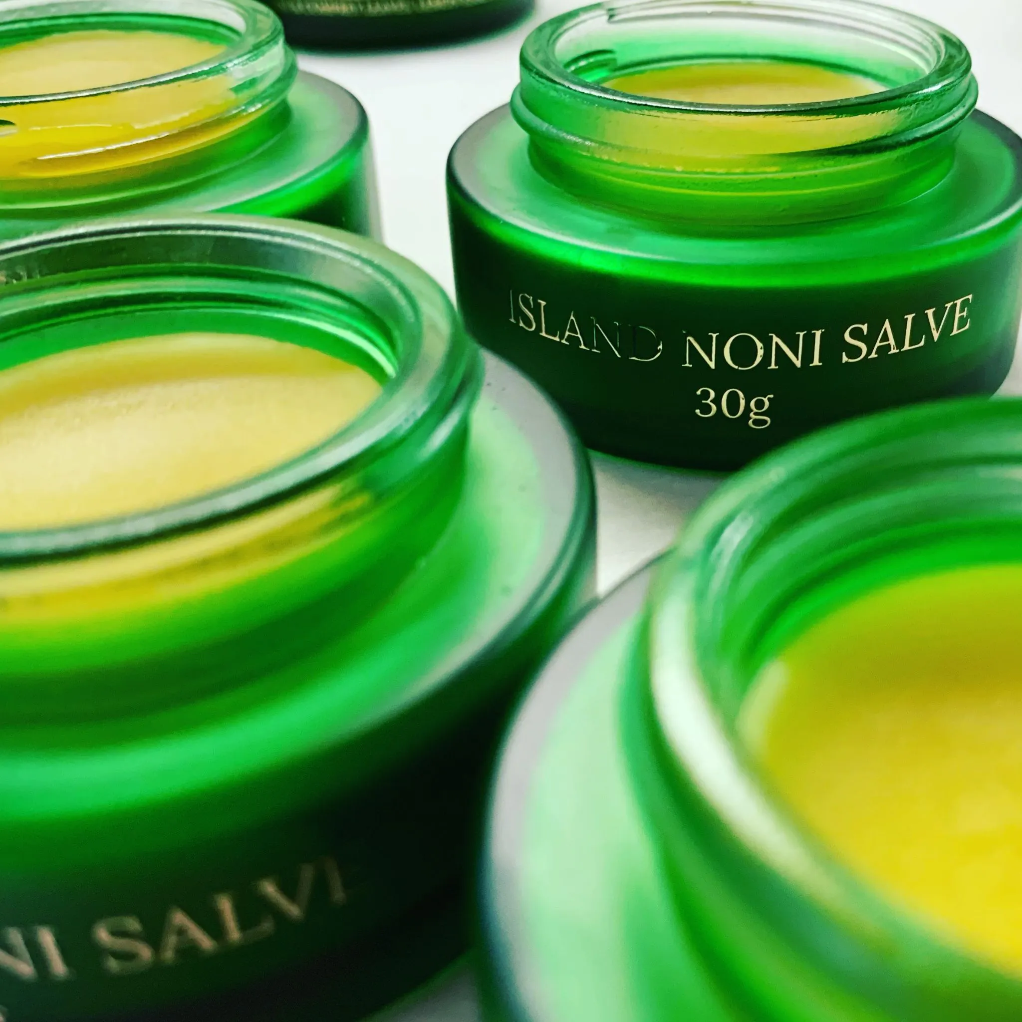 Close-up of several green glass jars filled with golden Maui CBD Infusions Island Noni Salve.